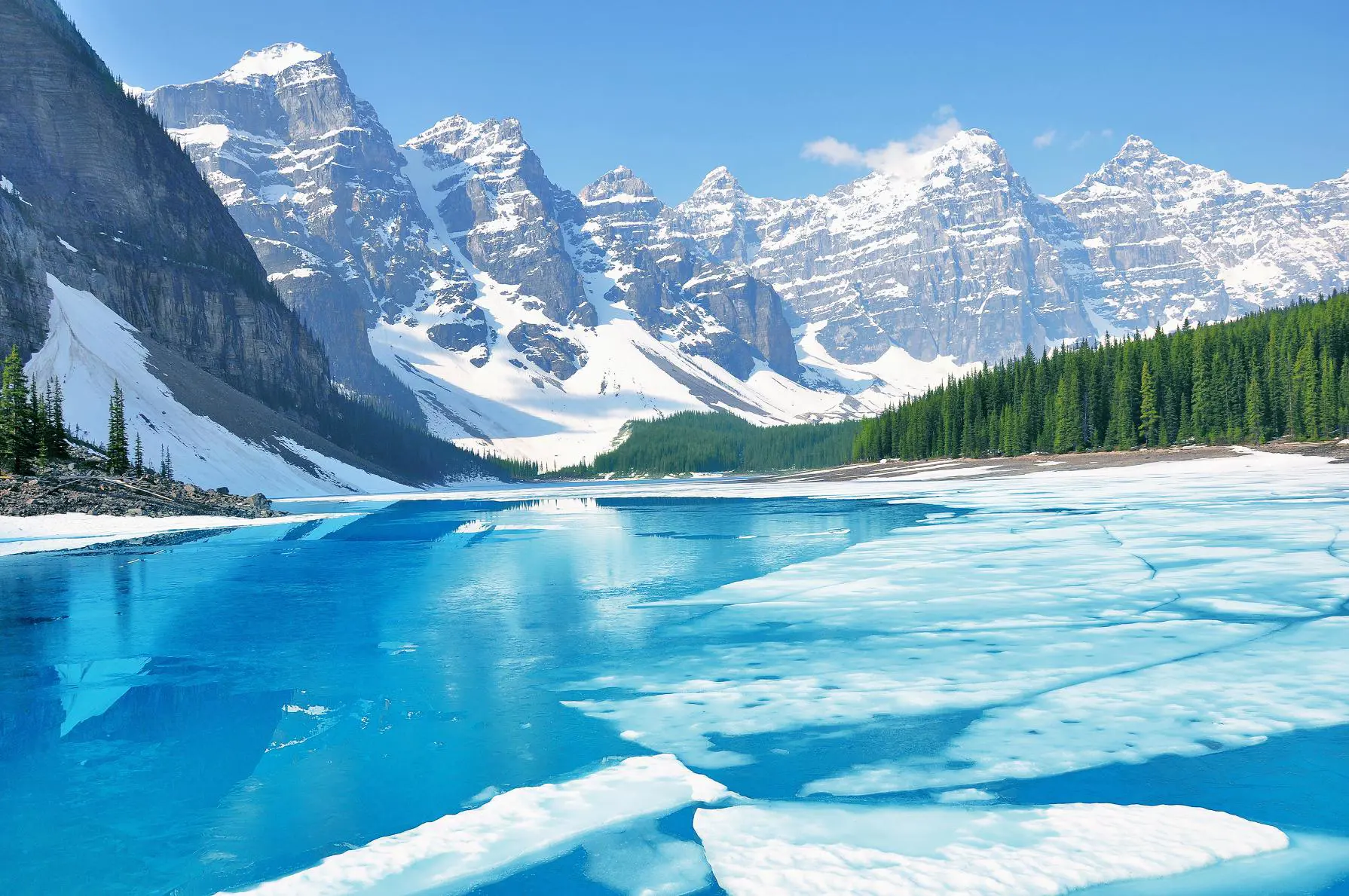 Moraine Lake partially covered by ice in the early morning during spring, with soft light illuminating the frozen surface and surrounding mountains in Banff National Park, Canad