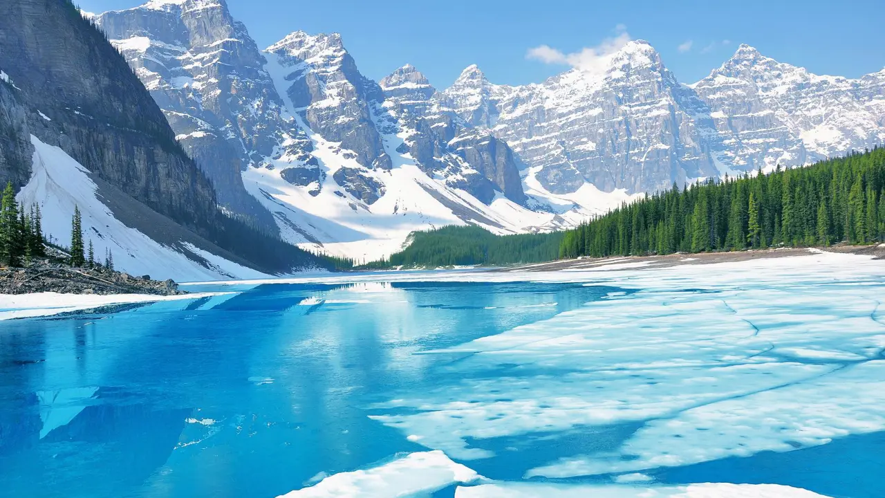 Moraine Lake partially covered by ice in the early morning during spring, with soft light illuminating the frozen surface and surrounding mountains in Banff National Park, Canad