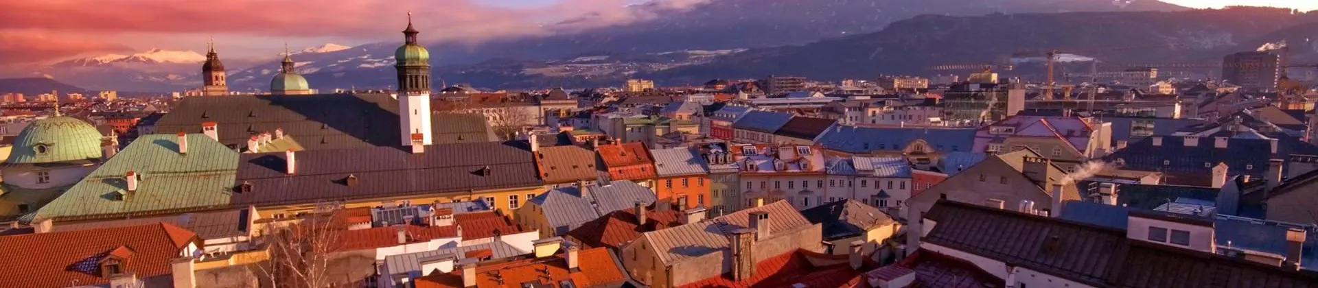 High angle shot of the city of Innsbruck, showing many roofs of houses and other buildings, with a few towers poking out, and one with a dome-shaped roof. Large mountains in the distance, dark pink clouds to the left and a blue sky. 