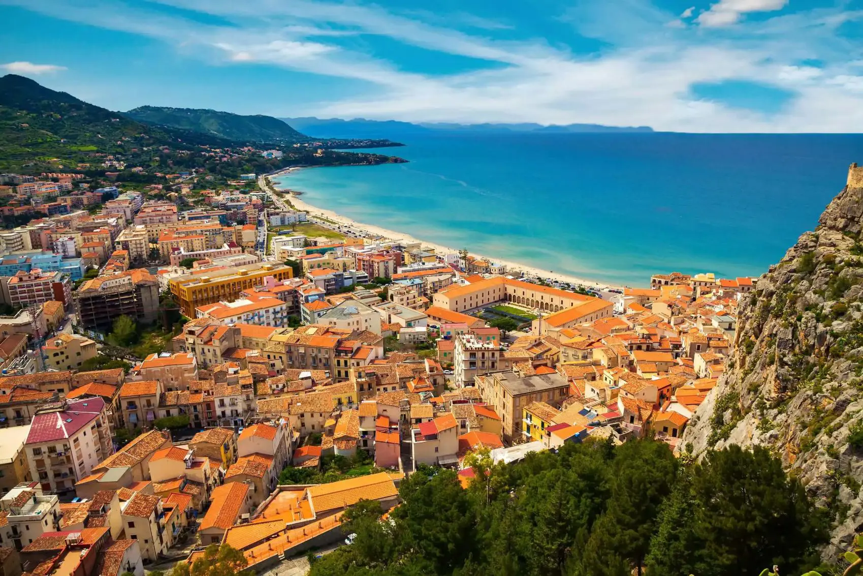 A panoramic view of Cefalù, Sicily, featuring its historic seaside town with terracotta rooftops, a sandy beach, and the rugged coastline beneath a lightly cloudy sky