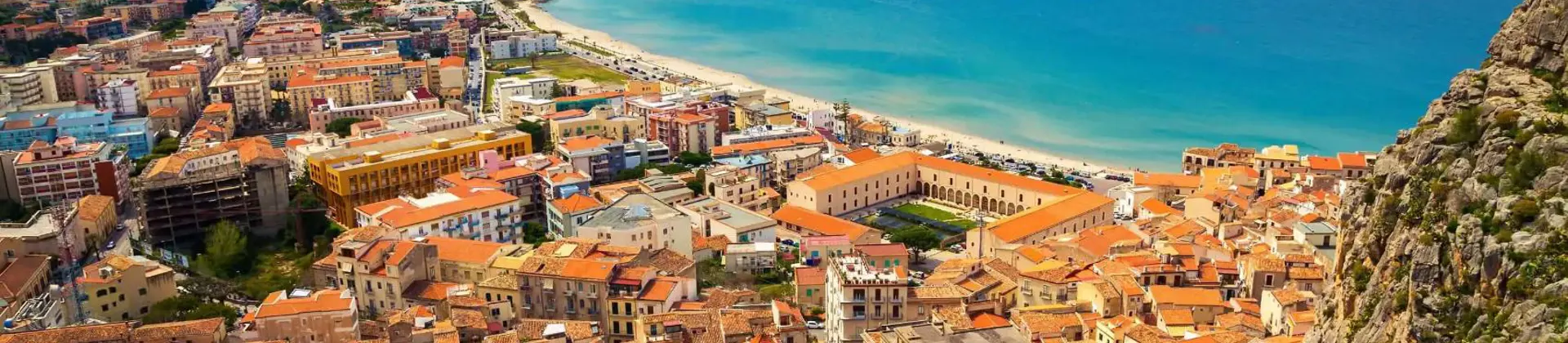 A panoramic view of Cefalù, Sicily, featuring its historic seaside town with terracotta rooftops, a sandy beach, and the rugged coastline beneath a lightly cloudy sky