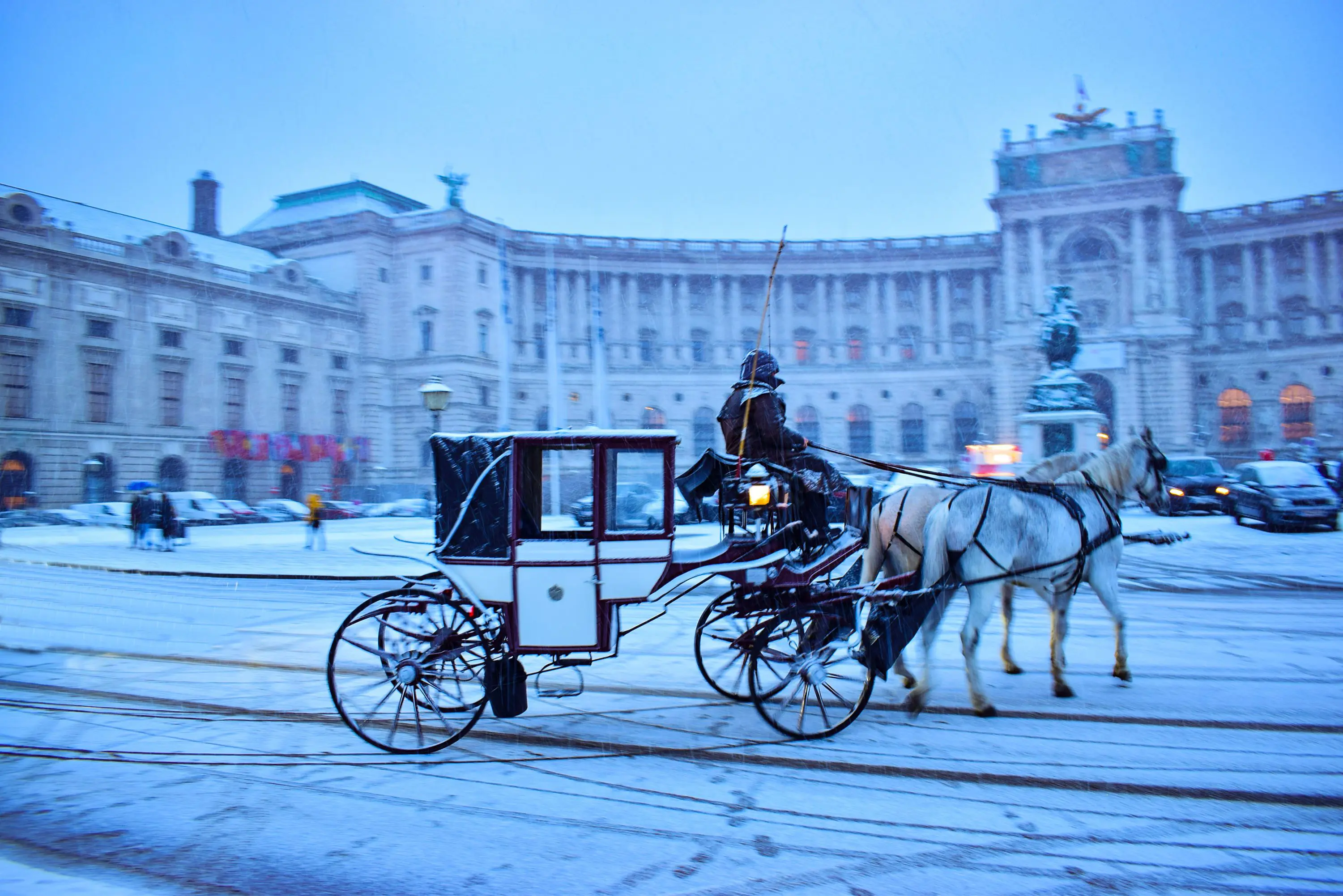 Action shot of a horse drawn carriage outside a large, wide, beige-coloured historical building, which also has a black statue of a person outside the building. Cars can be seen parked to the left of the image, and some moving on the right. Everything in the image is covered in snow