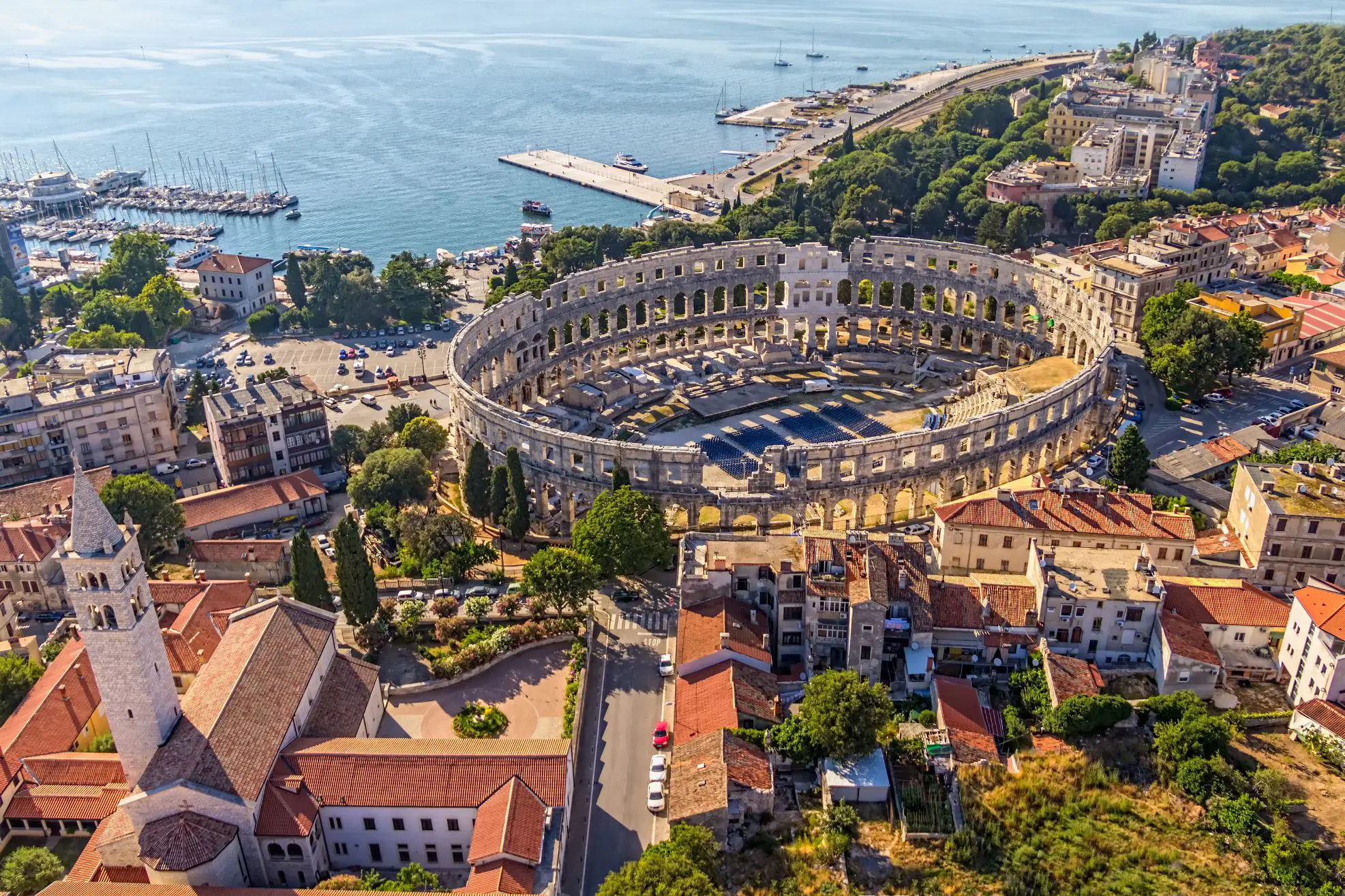 Bird's eye view of the ruins of an Amphitheatre and its surrounding area. A church with a tower in the left forefront, and regular buildings to the right. A boat harbour to the top right of the image, next to the rest of the sea to the right. 