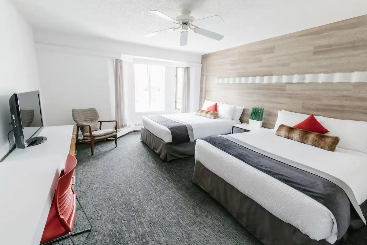 Spacious guestroom at Elk + Avenue hotel with two double beds, white bedding, grey bed skirts, a ceiling fan, a desk with a red chair, a flat-screen TV, and large windows allowing natural light to flood in