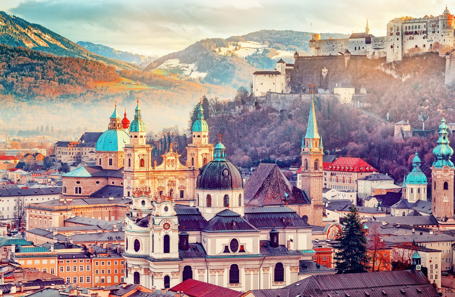Shot of Salzburg, showing the top half of the city's buildings, with turquoise turrets dotted around the image and a wide church in the forefront and a cathedral behind it. Mountainous landscape behind and the fortress on the hill to the right.