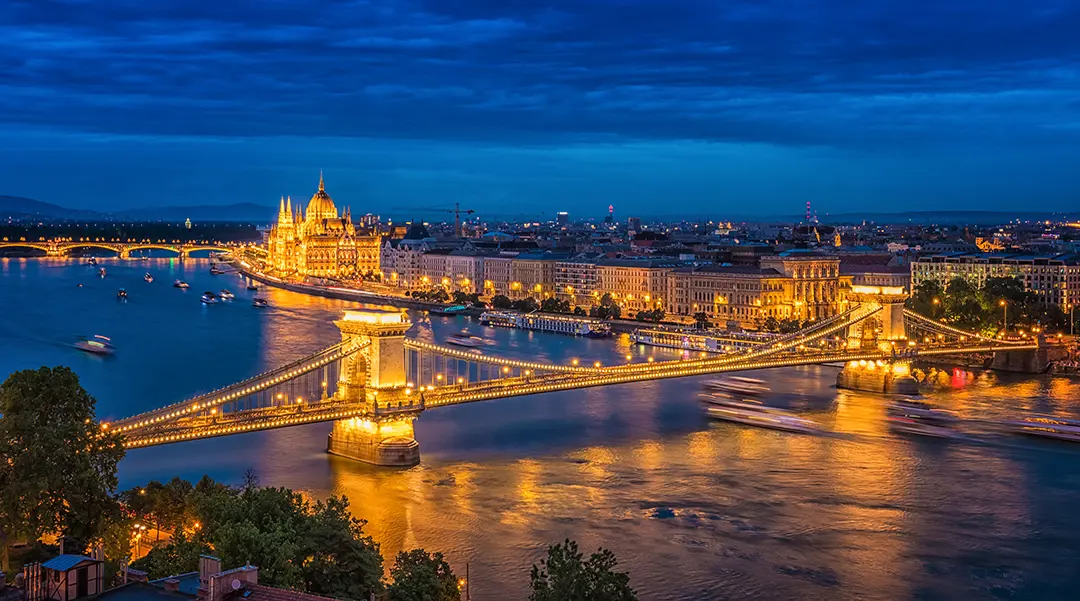High angle shot of Széchenyi Chain Bridge, going over the Danube River at night time. With a view of the Pest side, the Parliament building in the distance