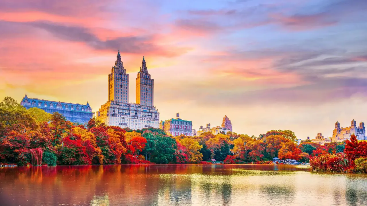 Central Park in autumn with The Lake in the centre, surrounded by colourful trees in shades of red, orange, green and yellow. The iconic twin towers of The San Remo rise in the background