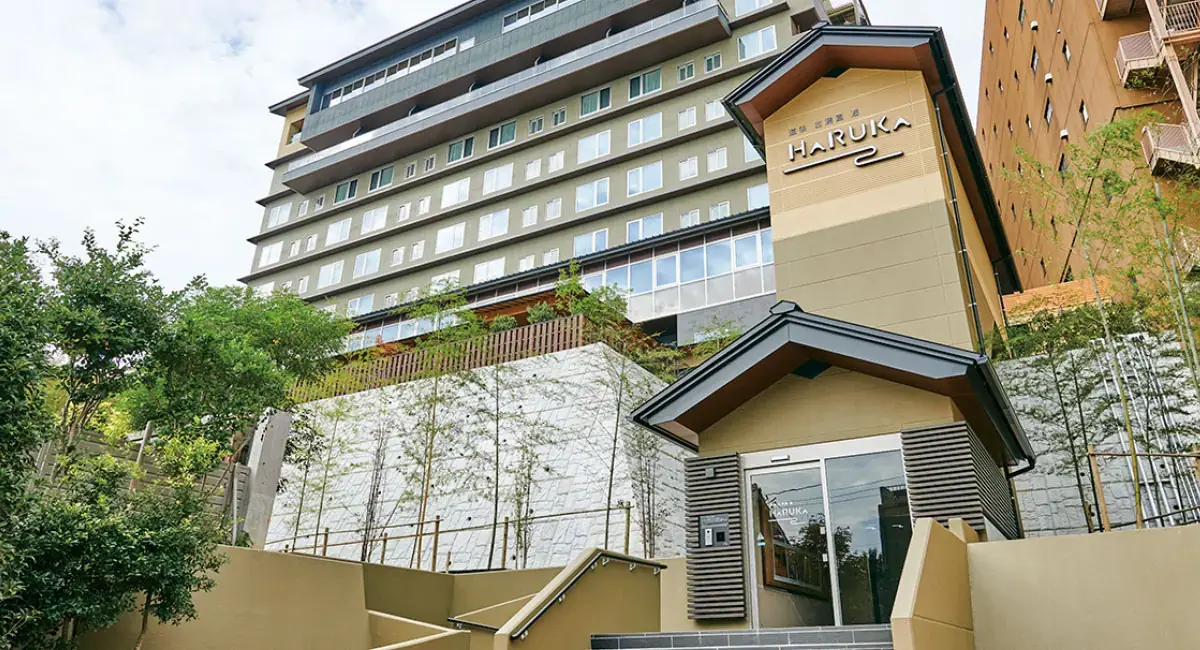 Exterior of the Hotel Kowakuen Haruka in Matsuyama, a multi-storey building nestled on a hillside with landscaped greenery and bamboo in the foreground