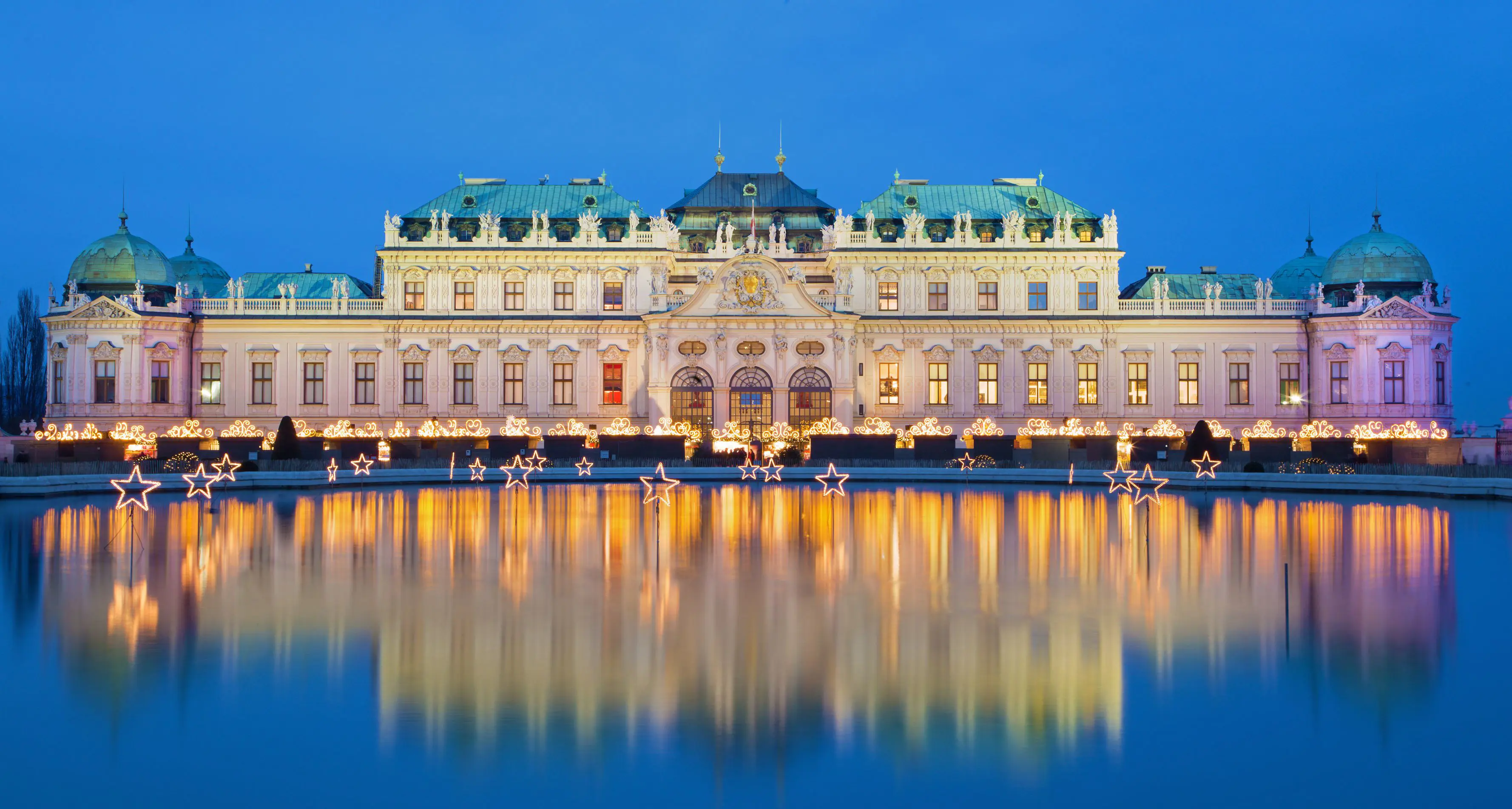 Shot of Schoenbrunn Palace, a wide, beige coloured building with turquoise roofs, in front of a bright blue night sky. The palace has gold, swirly light fixtures along the bottom. In front, is a large pond with lit, stand up stars dotted in.