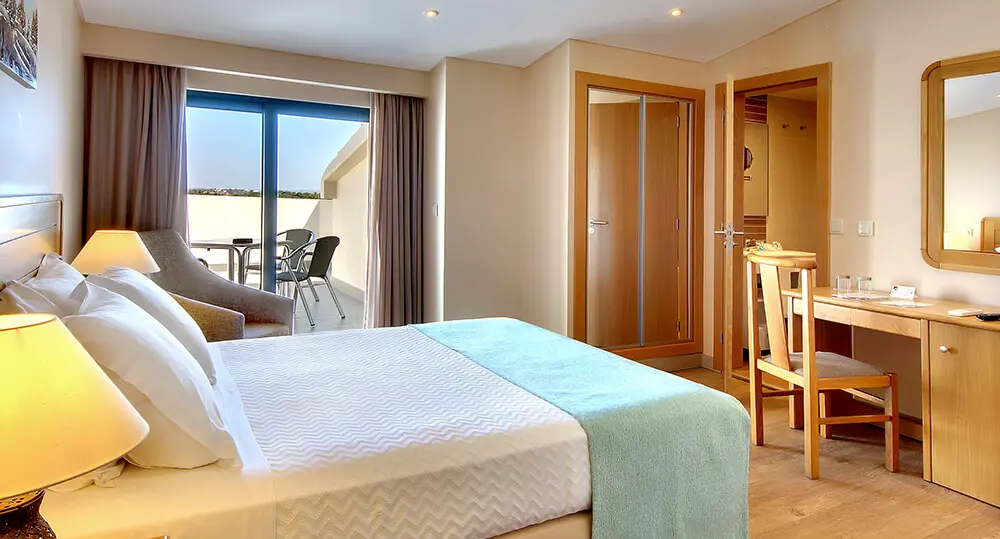 Modern double room at Hotel Alba in Monte Gordo, featuring crisp white linens, a blue bed runner, and wooden furnishings. A terrace with a dining table and chairs is accessed through a patio door