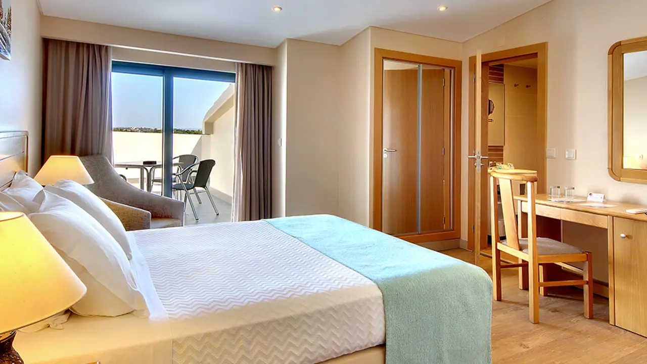 Modern double room at Hotel Alba in Monte Gordo, featuring crisp white linens, a blue bed runner, and wooden furnishings. A terrace with a dining table and chairs is accessed through a patio door