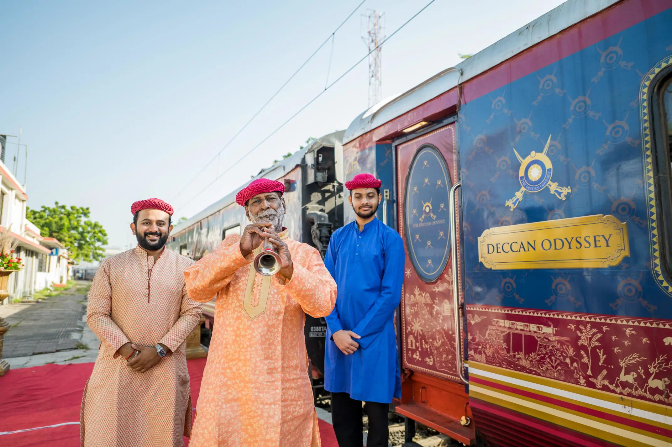 Three people dressed in traditional Indian clothing standing on the platform outside the Deccan Odyssey train, with the person in the middle playing a musical instrument