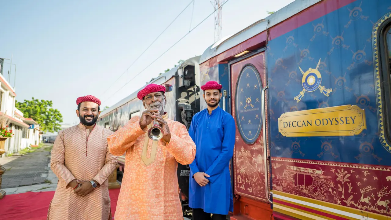 Three people dressed in traditional Indian clothing standing on the platform outside the Deccan Odyssey train, with the person in the middle playing a musical instrument