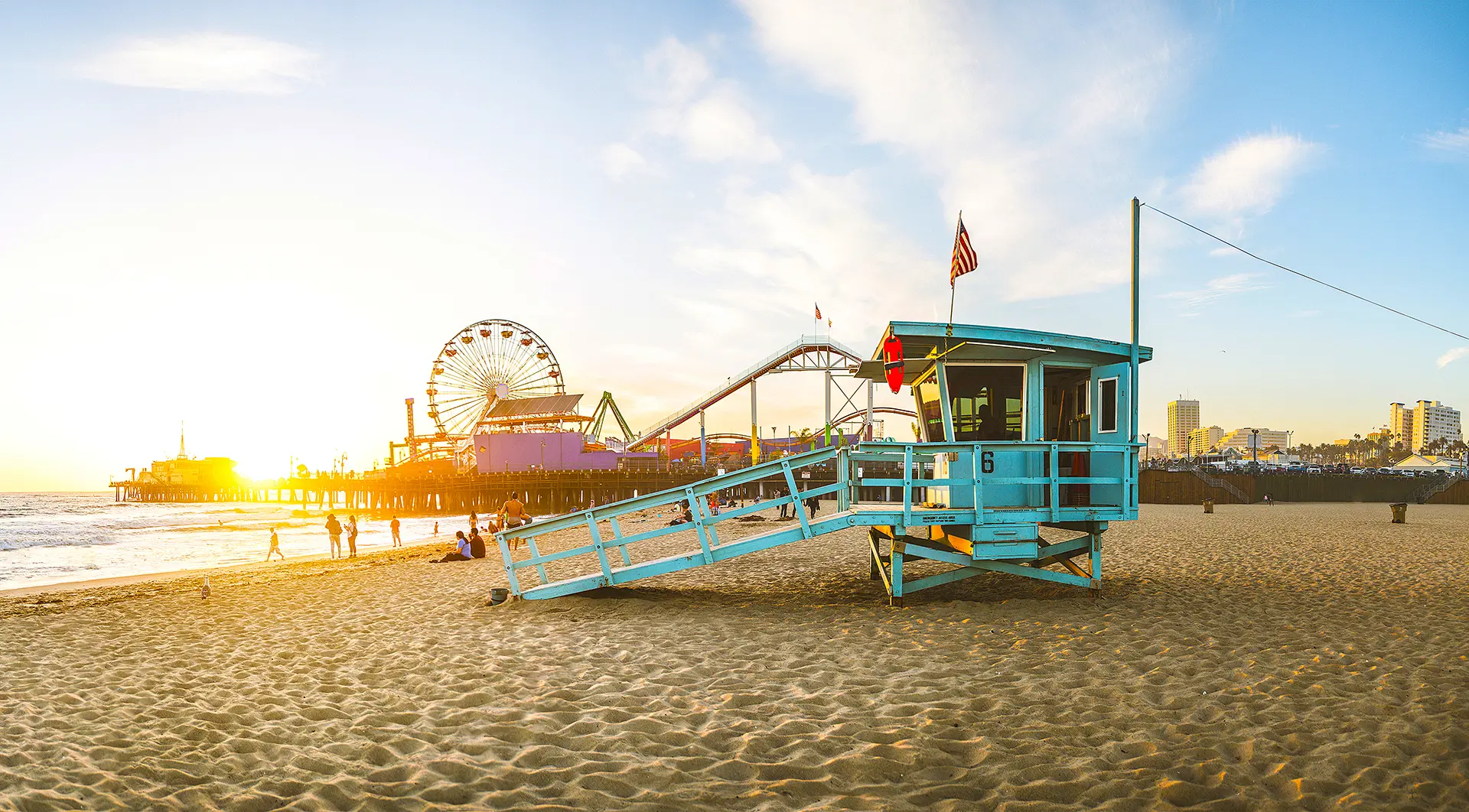 A teal blue lifeguard hut on Santa Monica Beach, with the iconic pier and Ferris wheel in the background as the sun goes down