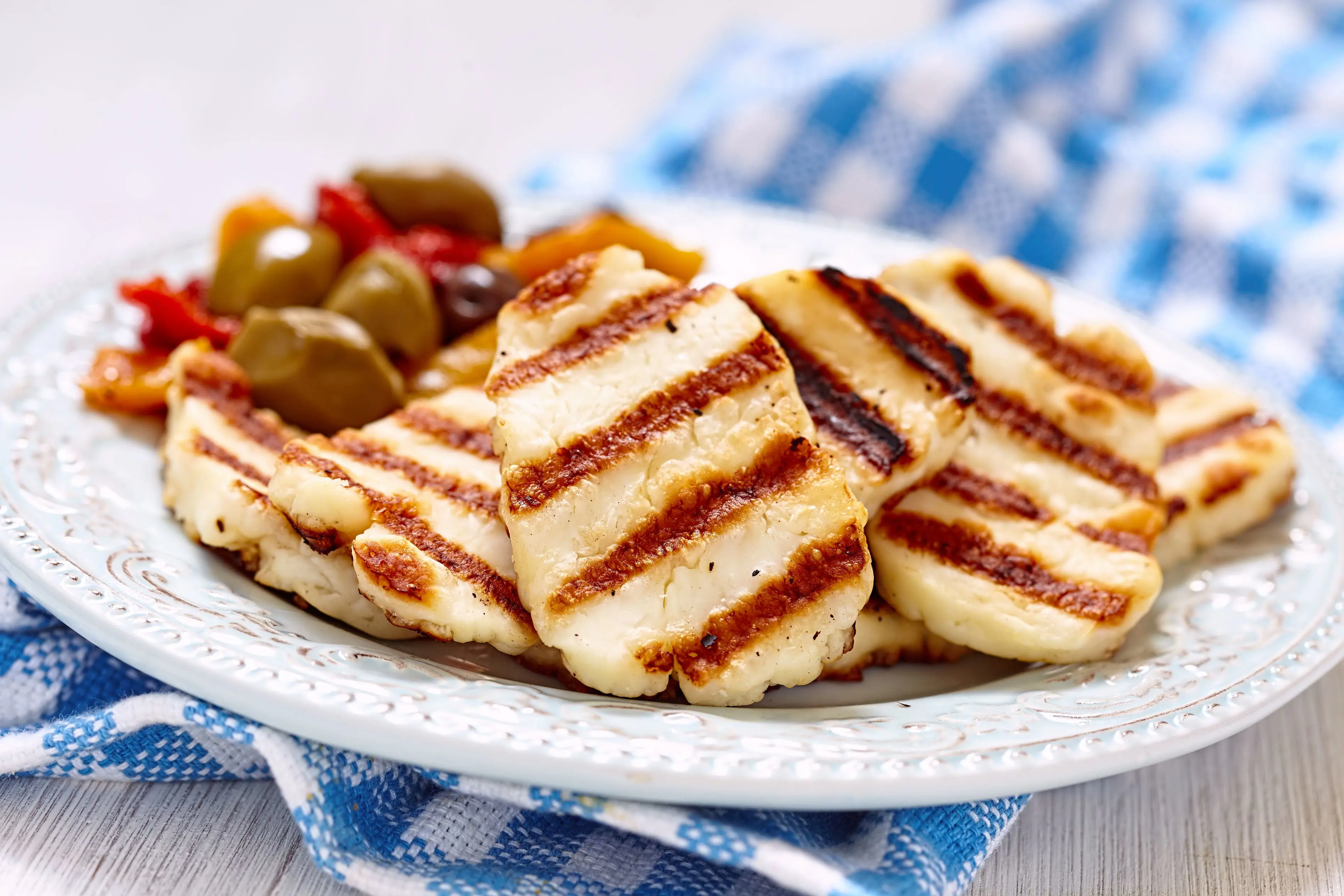 Close up of a dish of grilled halloumi with a side of olives, on a blue and white checkered cloth.