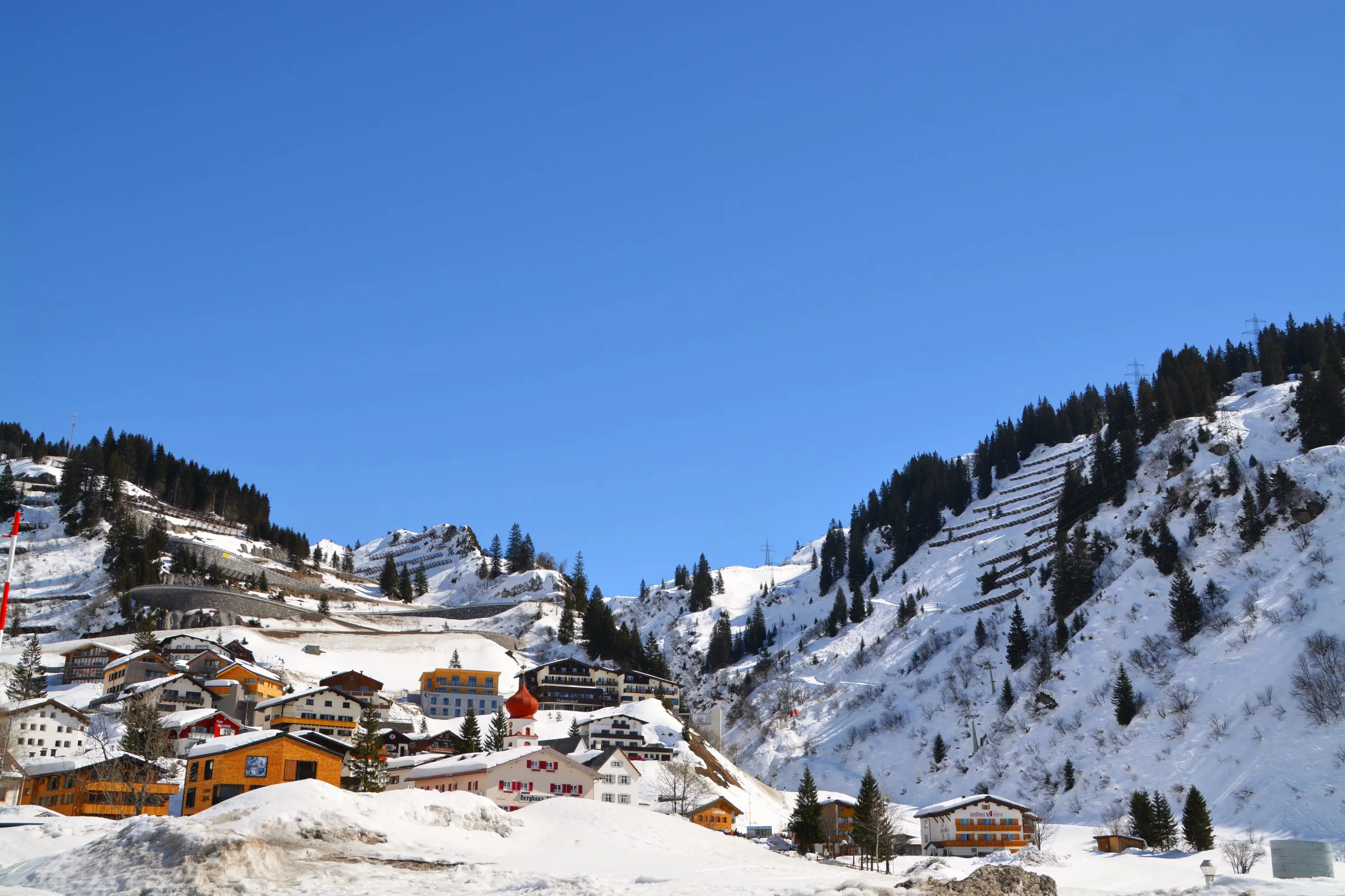 Low-angle view of St Anton village covered in thick snow, with a few houses and hotels going up the mountain and fir trees dotted all over. In front of a blue sky.
