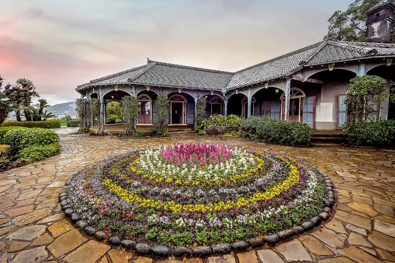 Historic Western-style buildings surrounded by lush greenery and colourful flowers at Glover Garden in Nagasaki, beneath a cloudy sky