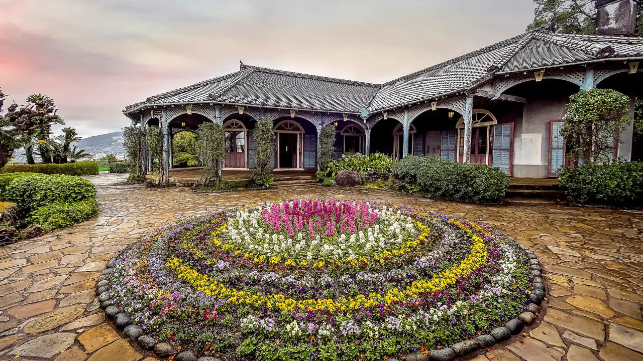 Historic Western-style buildings surrounded by lush greenery and colourful flowers at Glover Garden in Nagasaki, beneath a cloudy sky