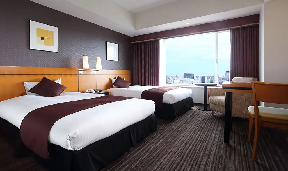 Contemporary twin hotel room at Hotel Nikko Ōita Oasis Tower, featuring two beds, large window with city views, and warm wooden furnishings