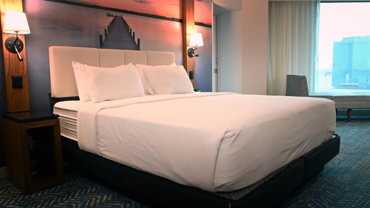 Modern king room at Fort Pontchartrain hotel, featuring a large bed with white linens, sunset-themed wall art, bedside lamps, a lounge chair, and a window with a city view