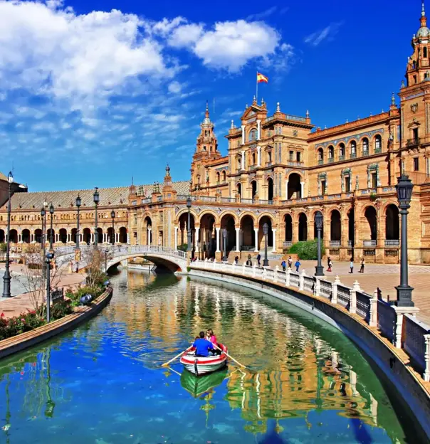 A couple rows a boat along the canal at Plaza de España in Seville, Spain, with the historic buildings and a bright blue sky reflected in the water