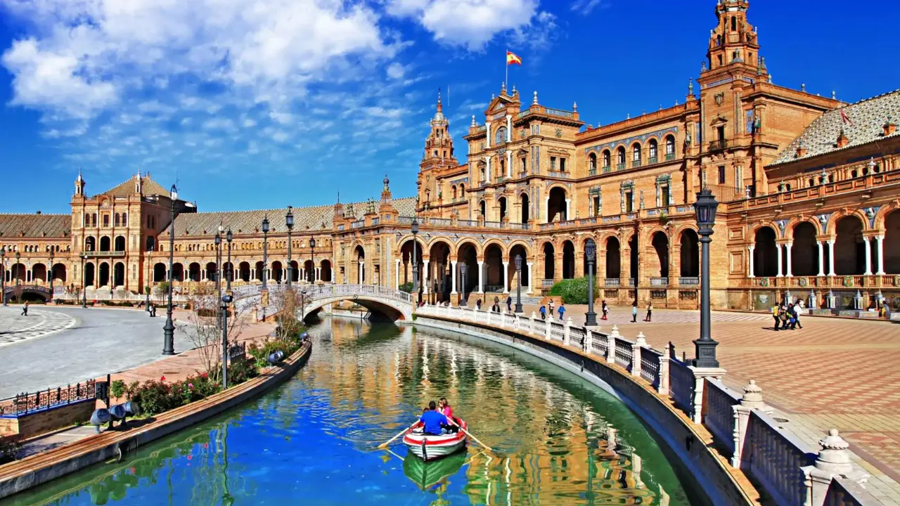 A couple rows a boat along the canal at Plaza de España in Seville, Spain, with the historic buildings and a bright blue sky reflected in the water