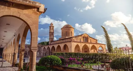A stone Greek Orthodox monastery with arched walkways, a domed roof, and a bell tower. A well-kept garden with flowers and shrubs lines the foreground under a sunny sky