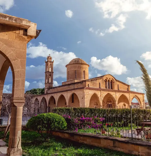 A stone Greek Orthodox monastery with arched walkways, a domed roof, and a bell tower. A well-kept garden with flowers and shrubs lines the foreground under a sunny sky