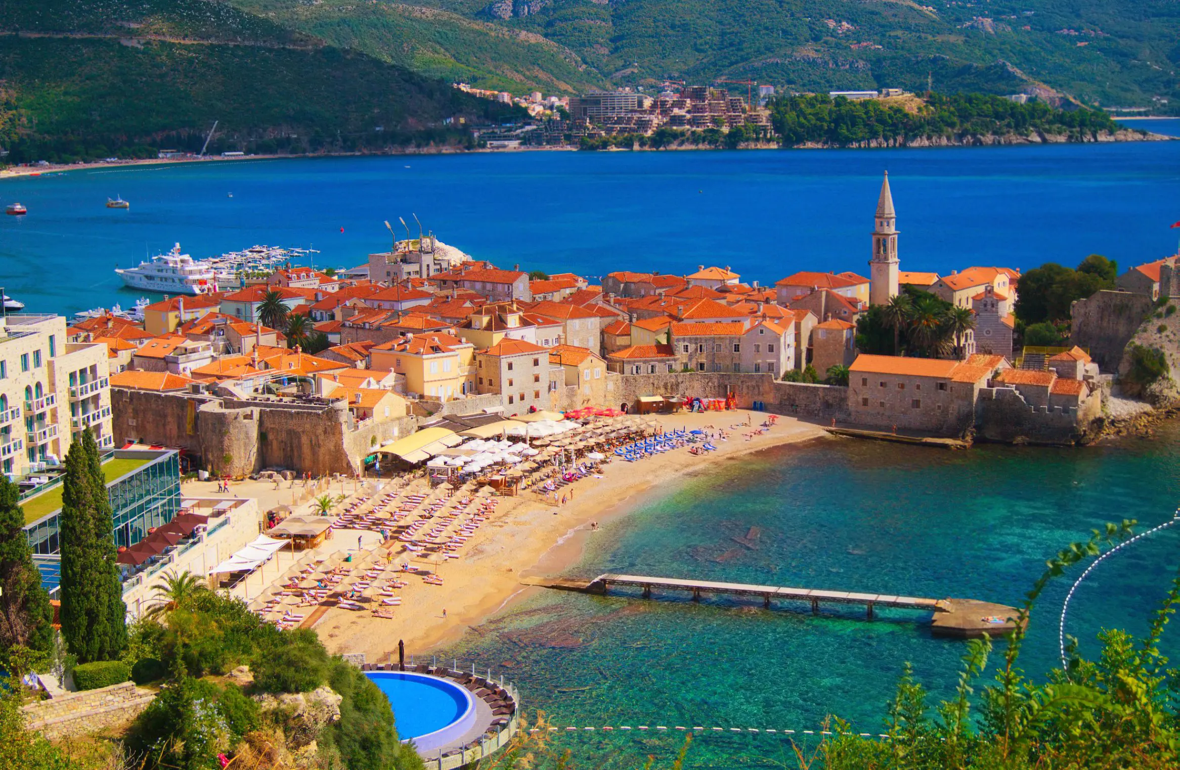  High angle view of a small beach, with sunloungers laid out and a turquoise sea. A group of buildings with orange roofs behind the beach and a bright blue sea behind there, with the start of a mountain behind it.