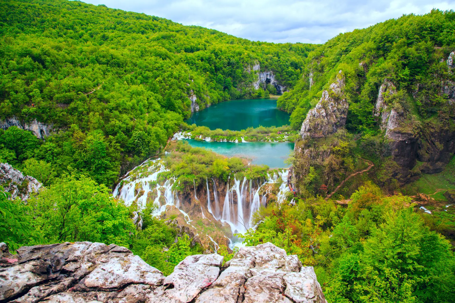 High shot of waterfalls in front of a dark turquoise body of water, surrounded by forested hills. In the forefront is a piece of rock and trees.