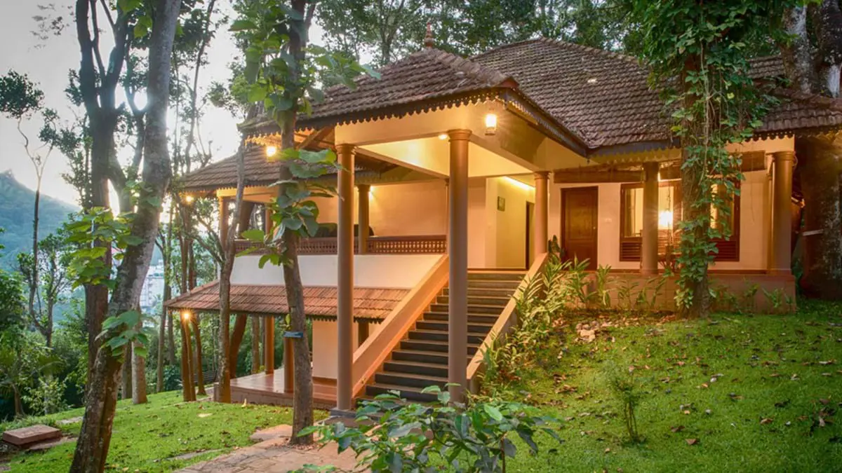 Exterior view of Greenwoods Hotel in Periyar, nestled among trees and greenery, with warm lighting highlighting the traditional building as dusk falls