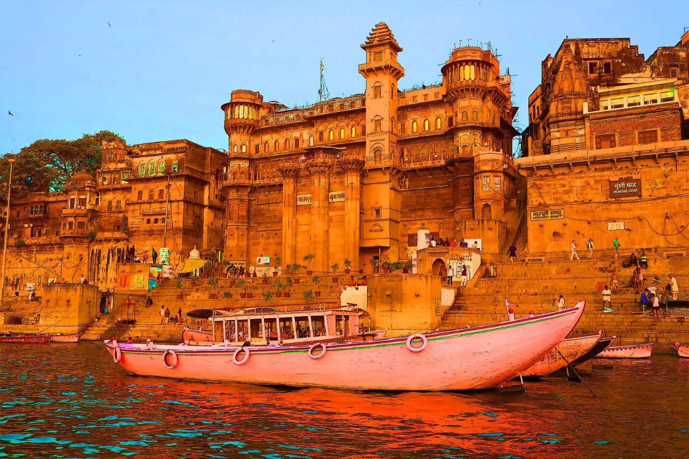 The ghats of Varanasi seen from the River Ganges, with historic sandstone palaces and colourful boats reflecting in the calm evening water