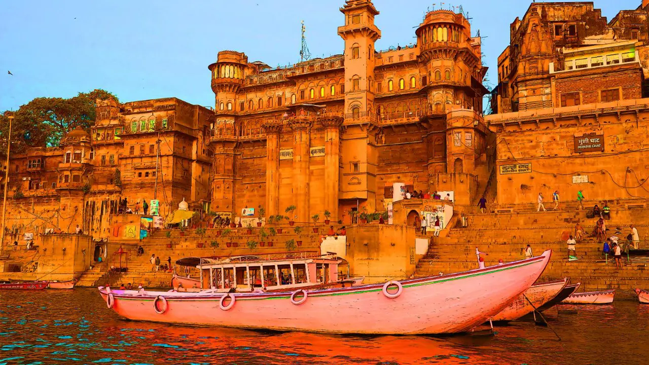 The ghats of Varanasi seen from the River Ganges, with historic sandstone palaces and colourful boats reflecting in the calm evening water