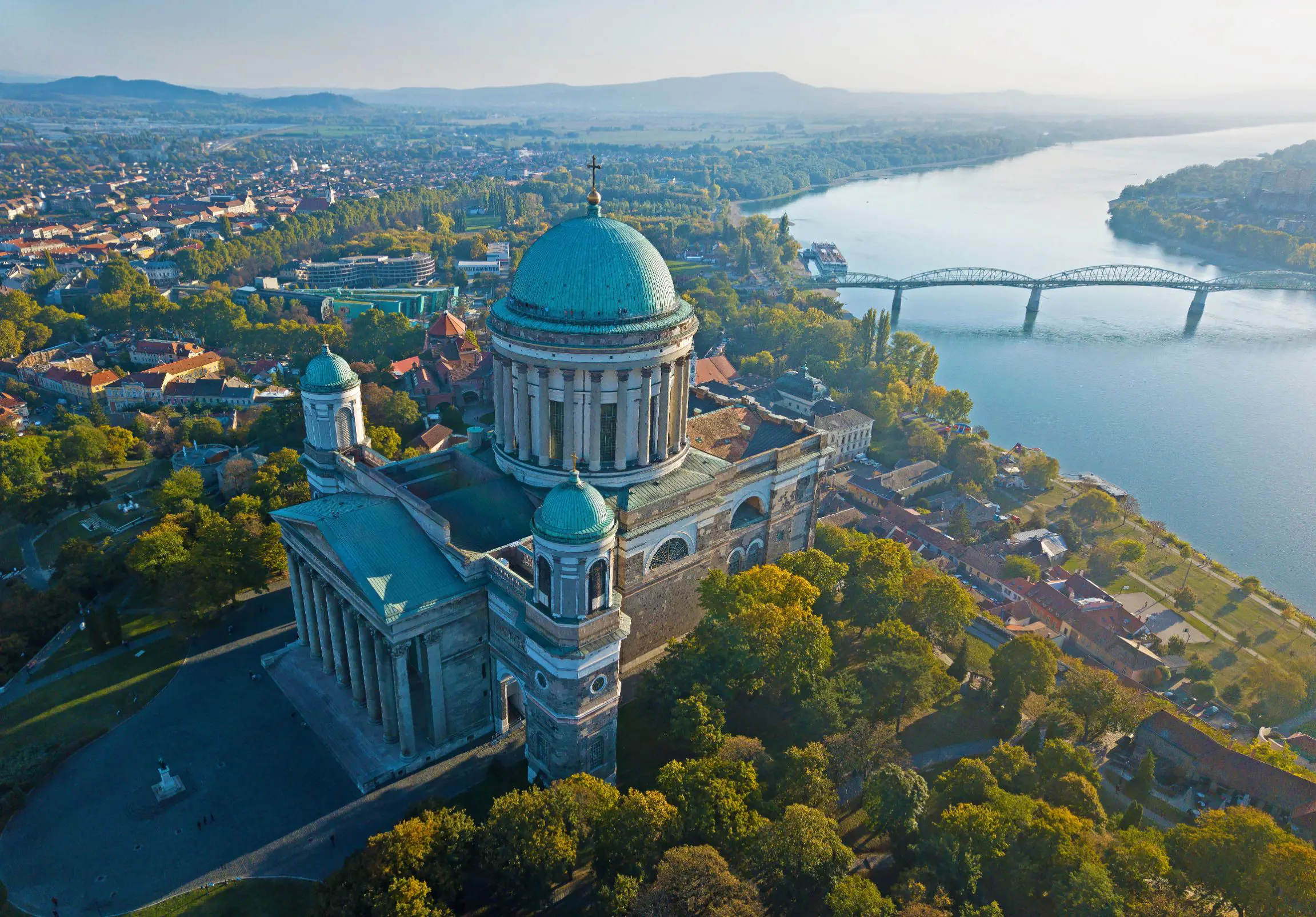 Bird's eye view of the Basilica of Esztergom, with it's turquoise domed roof with a cross on in the centre, and two smaller towers at the front with domed turquoise tops. Danube river in the background, with a bridge going across.
