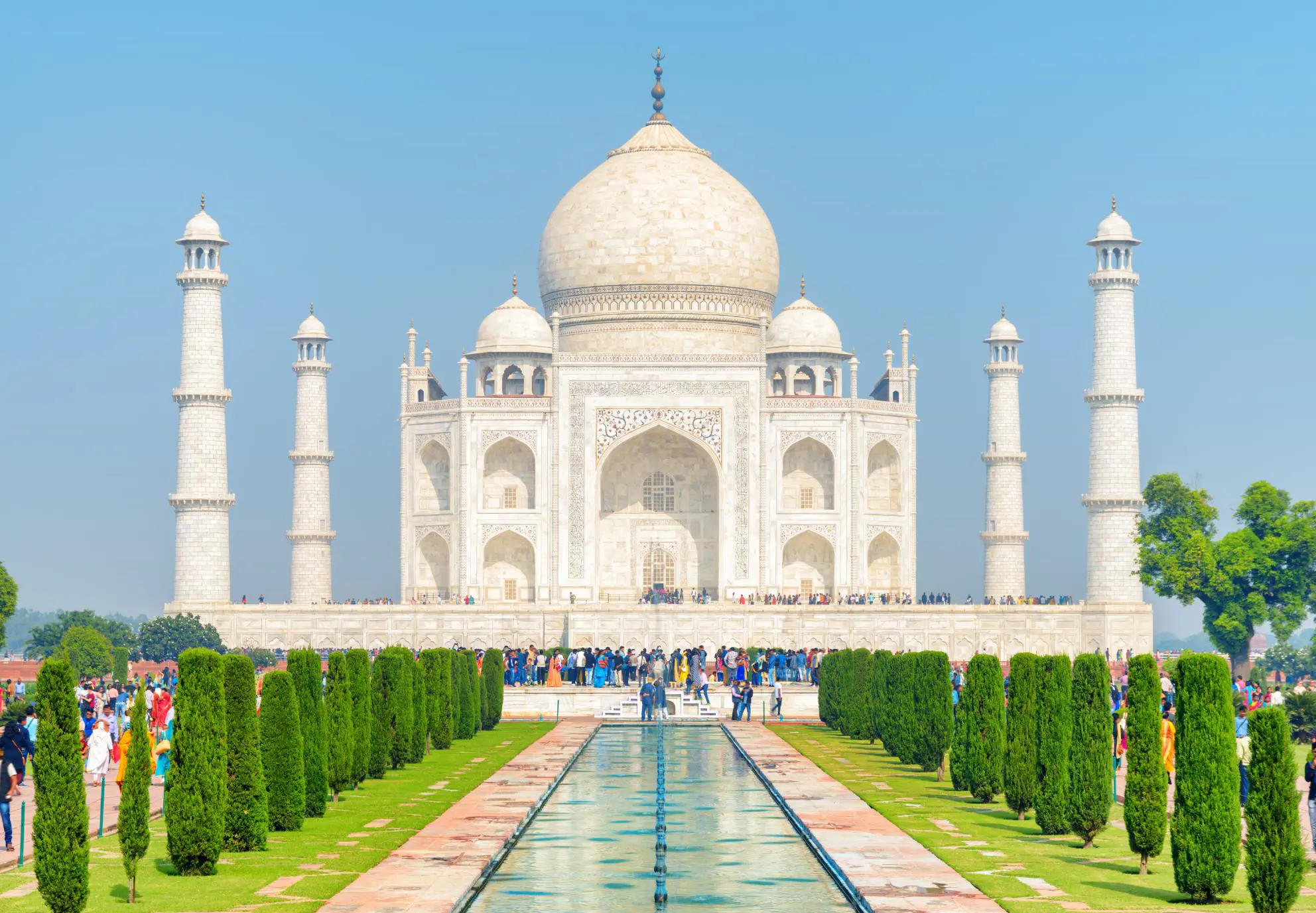 A symmetrical view of the Taj Mahal on a clear day, showing its white marble façade, ornate minarets, and carefully trimmed topiary gardens