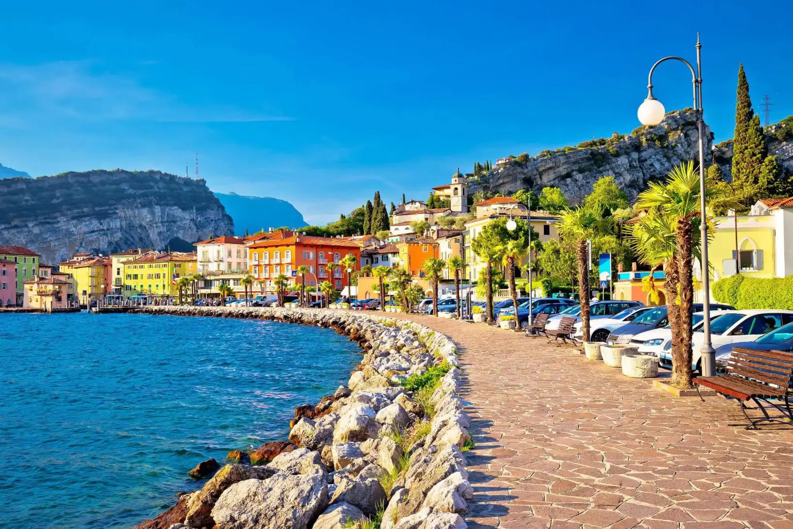 The waterfront in Torbole, Lago di Garda, featuring a paved promenade lined with colourful buildings and distant mountains rising beyond the lake