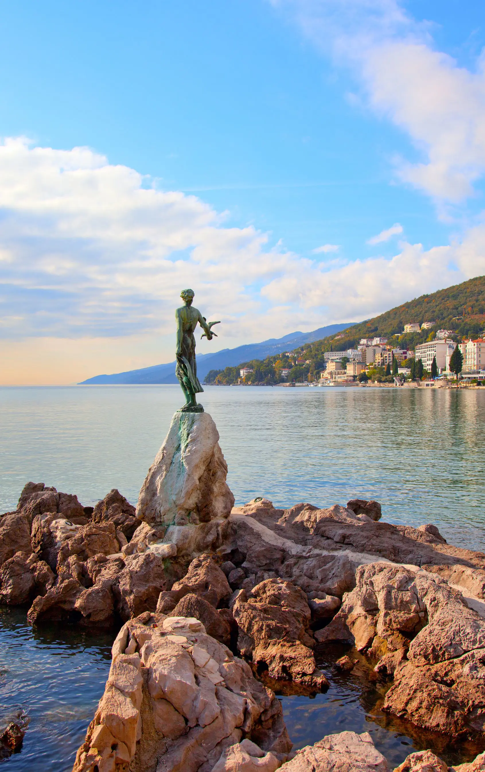 Back of a green statue of a lady holding a bird, on some rocks, facing the sea. Past the sea, there is a silhouette of a mountain, and on the right is a town. The sky is blue but cloudy